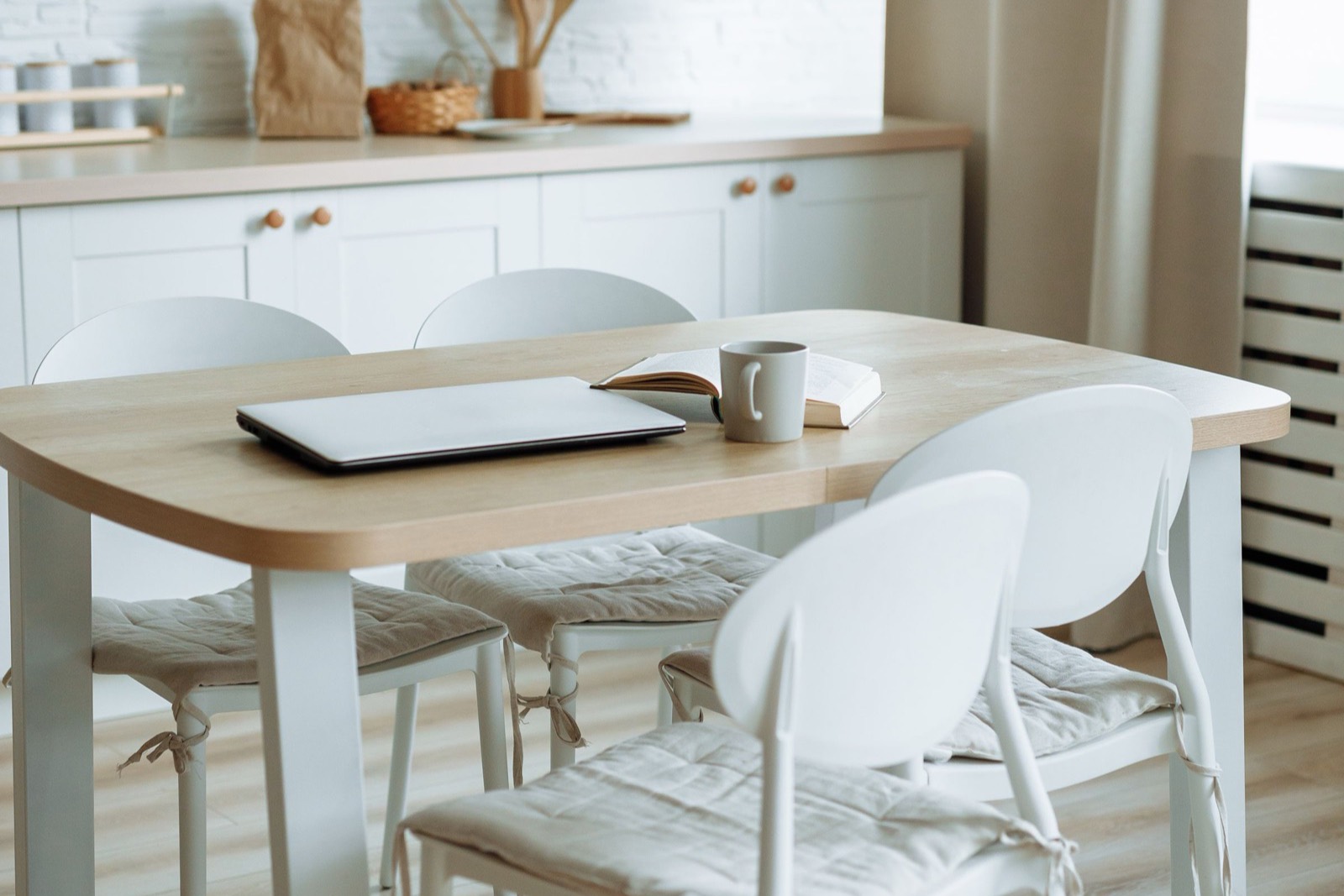 A laptop, a mug and an open book on a pale timber dining table in a bright kitchen, chairs pushed in around it.