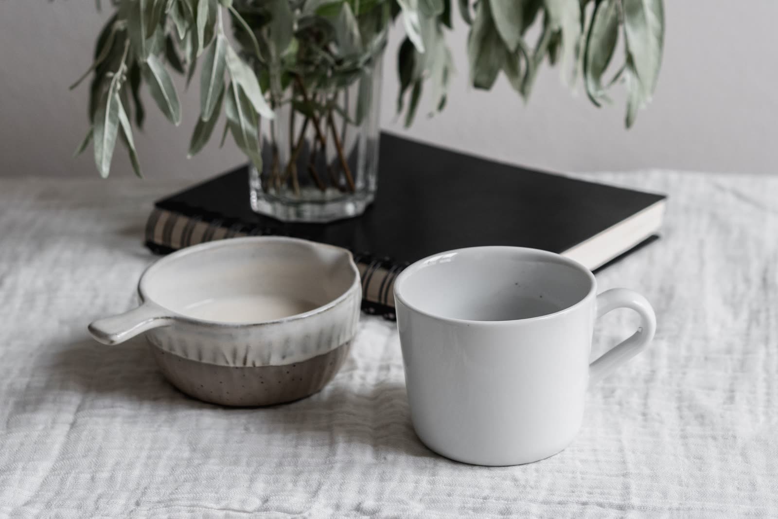 A white mug and a small ceramic jug beside a closed black notebook and a glass vase of olive foliage on a linen tablecloth.