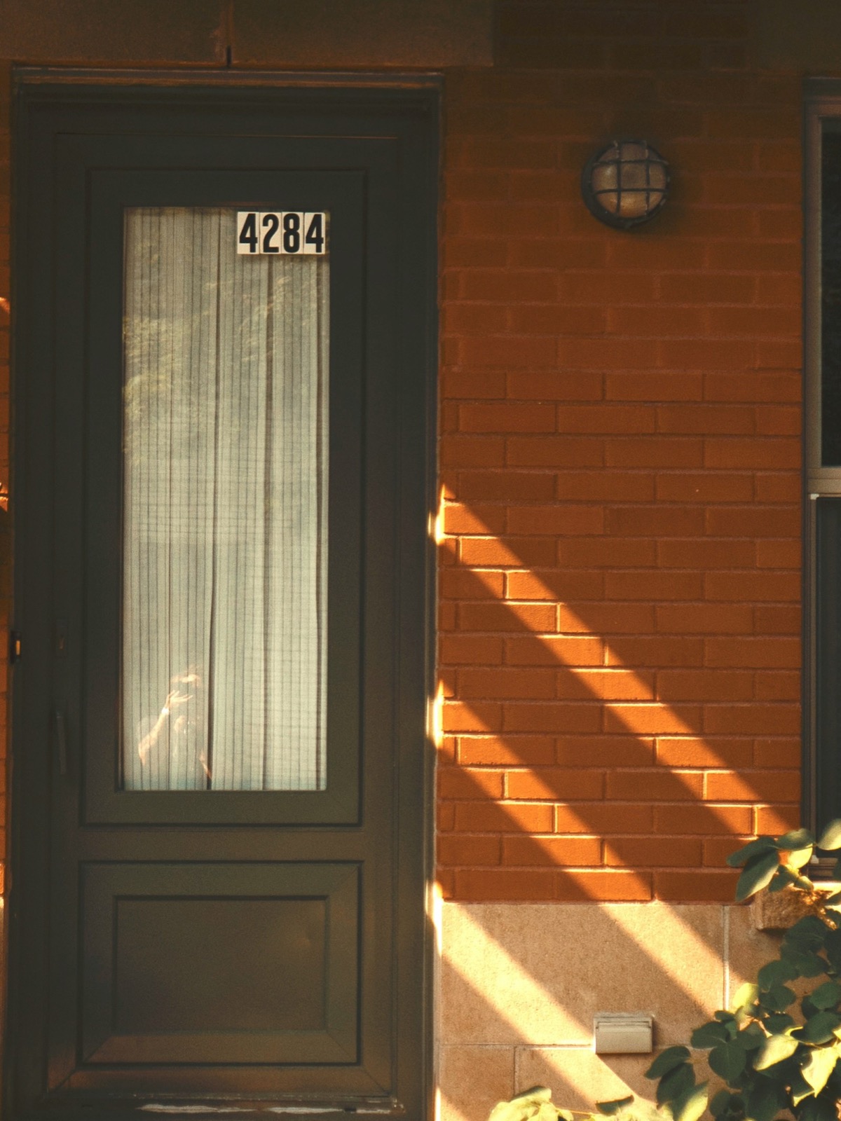 The front door of a brick house in late afternoon sun, a house number on the glazed panel and long shadows raking across the brickwork.