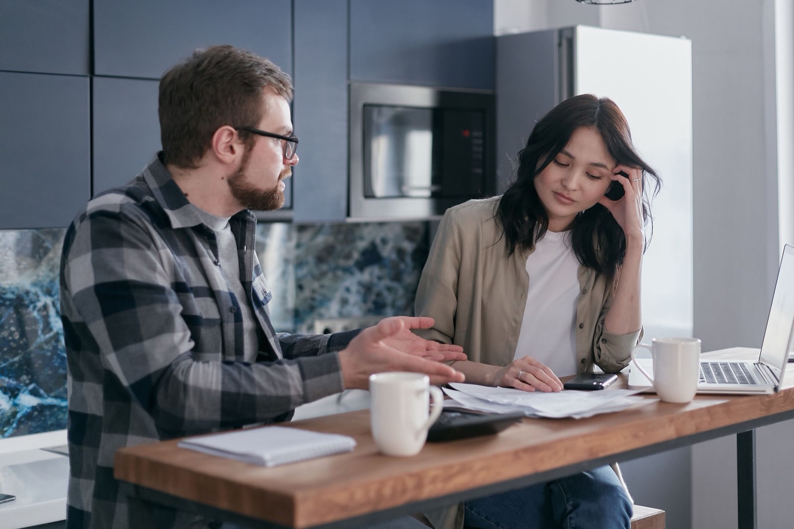Two people sitting at a timber kitchen bench, both turned away from the camera, with loose papers, a laptop, a notebook and two mugs between them.