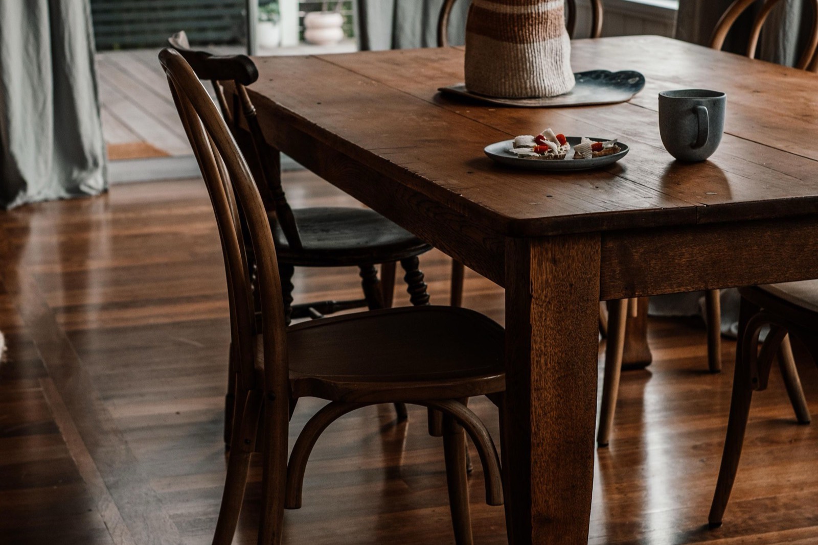 An empty timber dining table with bentwood chairs drawn up to it, a mug and a small plate on the tabletop, late afternoon light across polished floorboards.